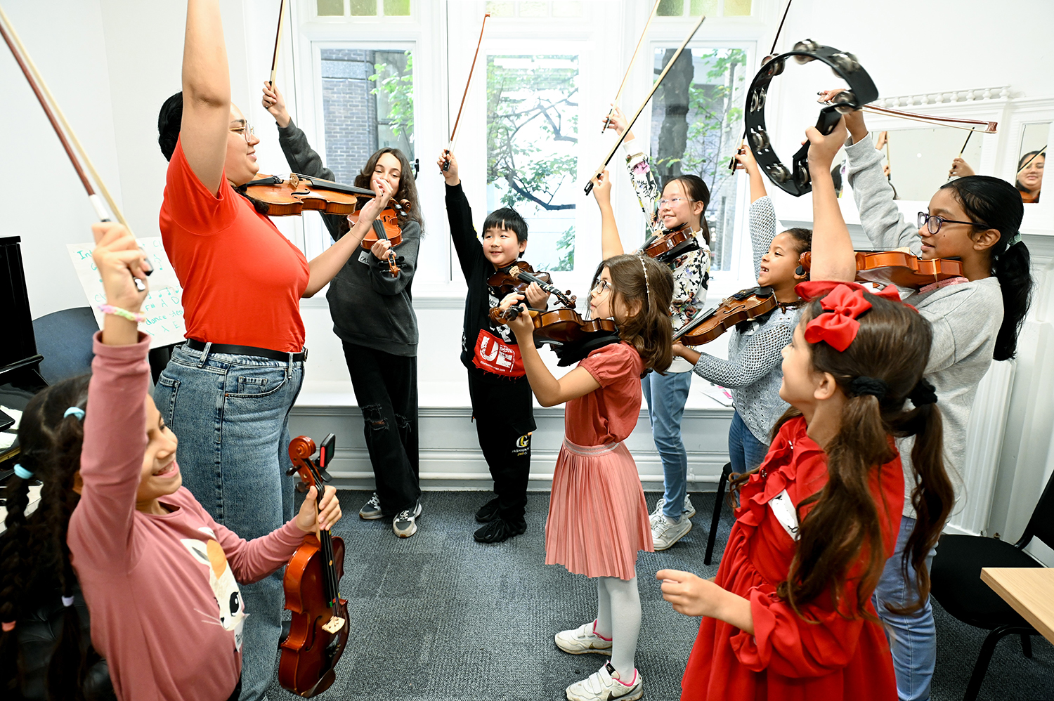 Children holding bows of a violin and tambourines in the air with an RCM Sparks member